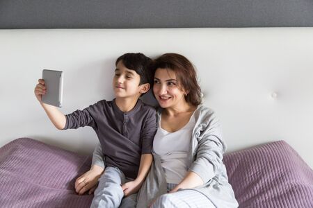 Mother and son having happy family time at home watching movie on tablet, taking a selfie, enjoying...