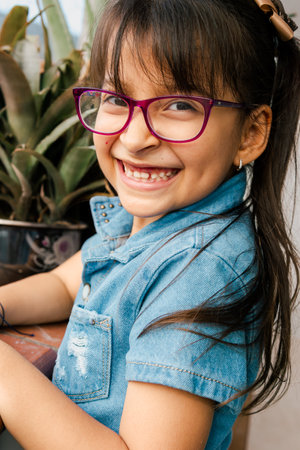 Beautiful Latina girl with pink glasses, smiling on the balcony of her house. Children day, family day. Infant and plants.の写真素材