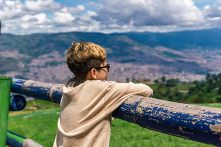 Colombian boy leaning on a rustic wooden fence, admiring a panoramic mountain view of a city under a bright and cloudy skyの写真素材