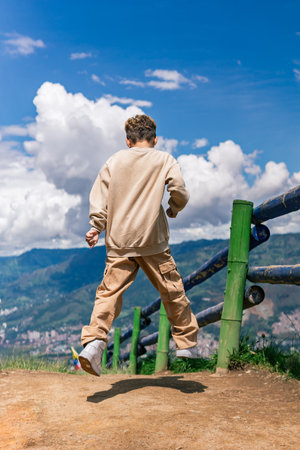 Latino boy jumping joyfully on a mountain trail under a sunny blue sky, full body rear view, wearing beige casual outfitの写真素材
