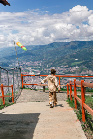 A young boy, approximately 11 years old, walks toward a scenic mountain overlook with a panoramic view of the city. Dressed in beige casual clothing, the child is captured from behind on a sunny day, surrounded by mountains, cityscape, and a colorful wind flag fluttering in the breeze. The image evokes a sense of adventure and peaceful exploration.の写真素材