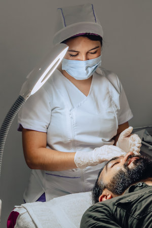 Photo of Colombian mestizo adult male with beard in a beauty spa with female dermatologist applying sunscreen on his face to complete a facial cleansing treatment.の写真素材