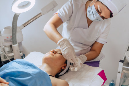 Photo of cosmetologist removing horse chestnut scrub from the face of a Colombian mestiza woman at a beauty spa where she is having a facial cleansing.の写真素材