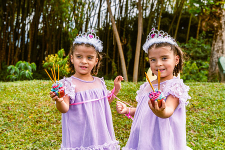 Colombian twin girls wearing tiaras and lavender dresses holding colorful cupcakes with long candles...