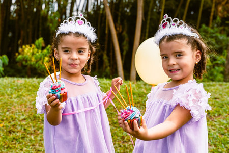 Colombian twin girls celebrating a birthday outdoors with tiaras, colorful cupcakes, and a yellow...