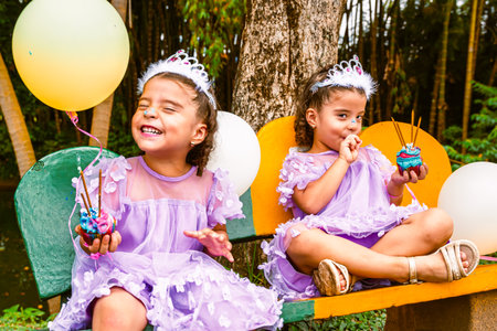 Twin sisters wearing tiaras and purple dresses holding cupcakes with candles and balloons while...