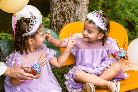 Colombian twin girls wearing tiaras and lavender dresses holding cupcakes with pretzels on a bench...