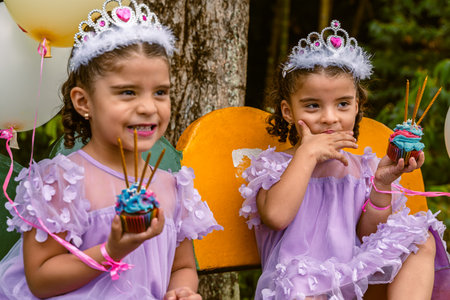 Twin girls wearing tiaras and lavender dresses holding blue cupcakes with candles on a yellow bench...