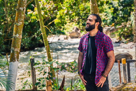 Smiling Colombian man with medium brown skin standing in tropical forest near river with copy-space...
