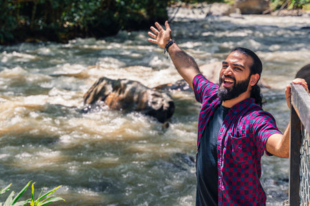 Colombian man with tied hair looking at camera while raising arm and holding fence near river in...