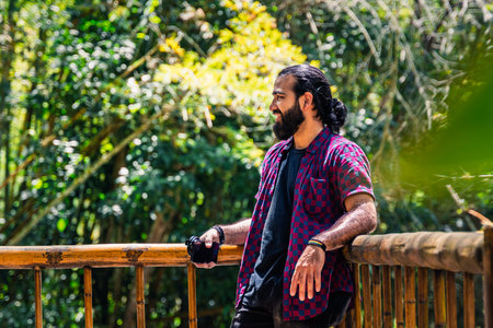 Man in plaid shirt with tied hair holding camera and gazing at tropical forest from wooden railing in Colombian jungle in Santuario, Risaralda, with space for textの写真素材