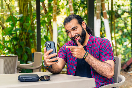 Expressive Colombian man in plaid shirt gesturing with phone during possible video call in lush tropical garden in Santuario, Risaralda, with space for textの写真素材