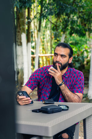 Man in early thirties sitting outdoors with focused expression, looking into distance while holding smartphone and contemplating a decisionの写真素材