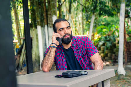Adult man sitting outdoors at a table surrounded by vegetation, possibly working remotely or enjoying a digital breakの写真素材