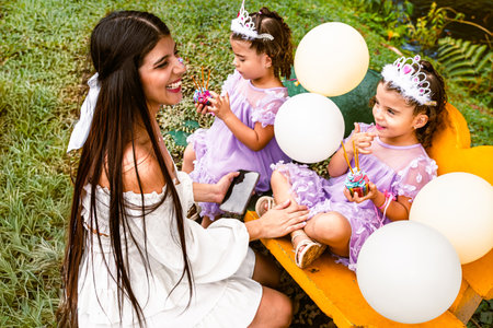 Colombian mother laughing with twin daughters in wheelbarrow during energetic birthday celebration...