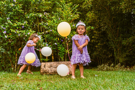Twin girls with tiaras and balloons play near log during outdoor birthday celebration