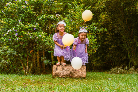 Twin girls with tiaras and balloons stand on tree stump surrounded by greenery during birthday...