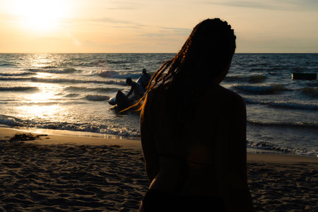 Silhouette of a Colombian woman with braids watching inflatable sea games at sunset on a beach near...