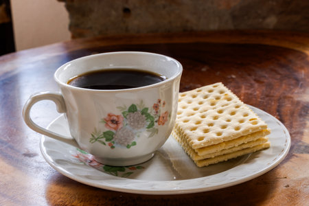 Cup of black coffee with floral design and stack of crackers on saucer, placed on rustic wooden...
