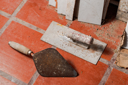 Pointed and plastering trowels on red tiled floor with stacked tiles and debris, showcasing essential masonry tools in realistic home renovation environment.の写真素材