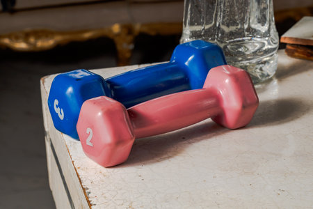Pink and blue dumbbells on cracked white surface with water glass and furniture detail, capturing...