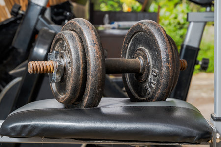 Rusty metal dumbbell with threaded collars resting on padded bench in home gym setting, featuring background equipment and natural light in strength training environment.の写真素材