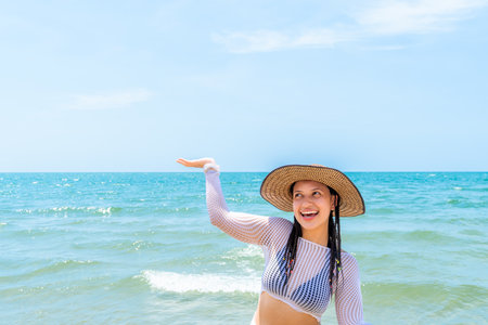 Smiling woman gestures toward the horizon under the sun, standing by the tropical beach with waves and sky behind her.の写真素材