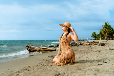 Caribbean woman kneels on the tropical beach in Covenhas wearing a handmade crochet cover-up, gazing calmly toward the sea under a soft coastal sky.の写真素材
