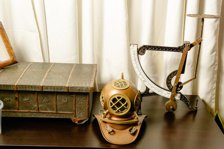 Vintage diving helmet, ornate chest and mechanical scale displayed on dark wood surface against white curtain in Colombian interior.の写真素材