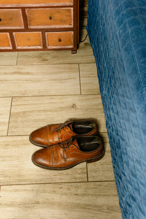 Brown leather dress shoes on wooden floor beside blue quilted bed and rustic dresser in Colombian bedroom.の写真素材