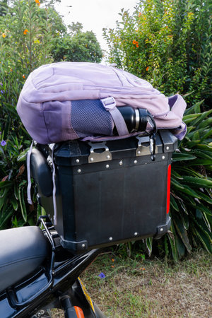 Purple backpack with water bottle and sunglasses resting on motorcycle top case in garden setting.の写真素材