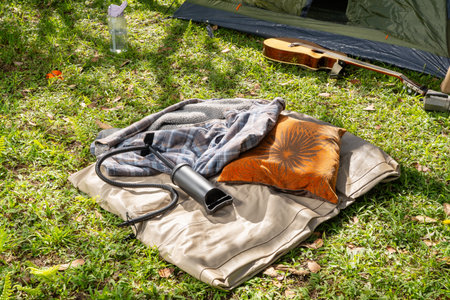 Camping setup with deflated mattress, blankets, pillow, tent, guitar, and water bottle on grassy...