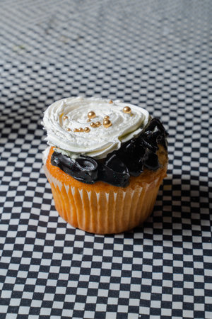 Cupcake with white and black frosting topped with gold beads on checkered surfaceの写真素材