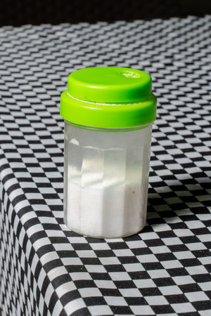Plastic container with green lid filled with white salt on checkered tableclothの写真素材