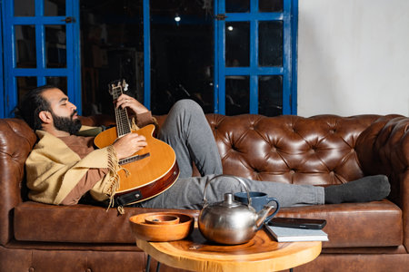 Person reclining with guitar and tea in cozy indoor setting with notebook and phoneの写真素材