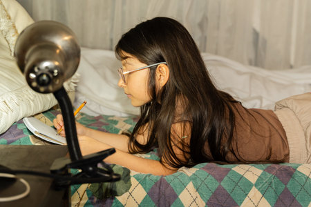 Young person studying on bed with notebook and lamp in cozy bedroom.の写真素材