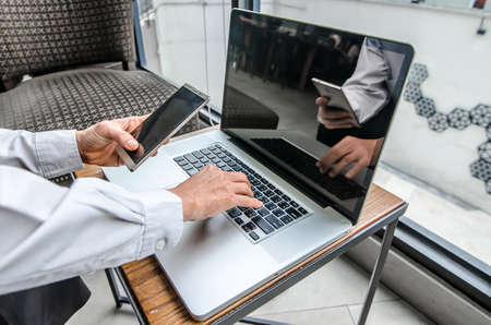 Portrait of young man with mobile phone working on laptop at indoor coffee shopの写真素材