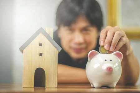 Man out of focus Inserting Coin In Piggy Bank of a wooden table: real estate, property investment, house mortgage, savings financial conceptの写真素材