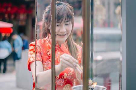 Cute chinese girl dressing traditional red Cheongsam suit burning incense sticks and pay respect and praying to Chinese God for luckyの写真素材