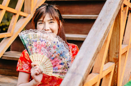 Portrait beautiful young woman smile wear cheongsam deep red dress holding a fan siting on stairs in...