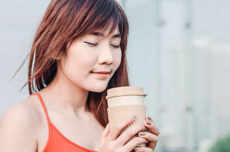 Close up beautiful young woman standing on balcony having cup of coffee, enjoying sun on a hot summer day. Asia female on terrace with city view in backgroundの写真素材