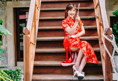 Portrait beautiful young woman smile wear cheongsam deep red dress holding a fan siting on stairs in sexy pose,in the Chinese New Year. Festivities, New Year Celebrationの写真素材