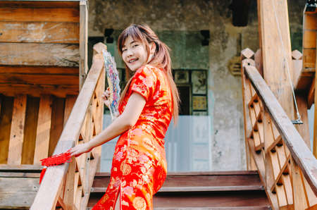 Portrait beautiful young woman smile wear cheongsam deep red dress holding a fan standing on stairs in sexy pose, Festivities and Celebration conceptの写真素材