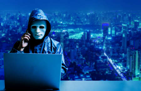 Anonymous computer hacker in white mask and hoodie. Obscured dark face using laptop computer for cyber attack and calling on cellphone  on modern city background, Data thief, internet attack, darknet and cyber security concept.の写真素材