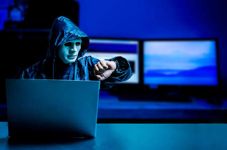 Anonymous computer hacker in white mask and hoodie. Obscured dark face using laptop computer for...