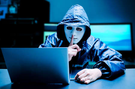 Anonymous computer hacker in white mask and hoodie. Obscured dark face making silence gesture try to...