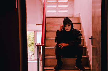 Portrait of white mask and hoodie sitting on old stairs. Selective focus on hands. Sadness, despair, dark, conceptの写真素材