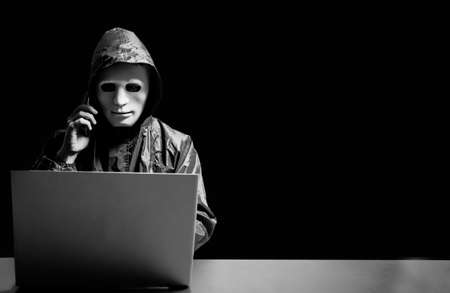Anonymous computer hacker in white mask and hoodie. Obscured dark face using laptop computer for...