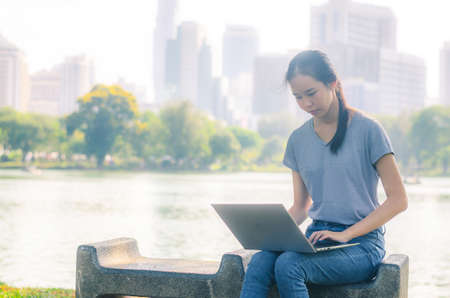 Portrait of pretty young woman sitting on bench  in park during summer day while using laptop of online shoppingの写真素材