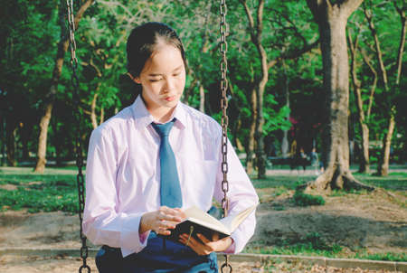 Lovely teenage girl wearing school uniform reading book while sitting on a swing at the park